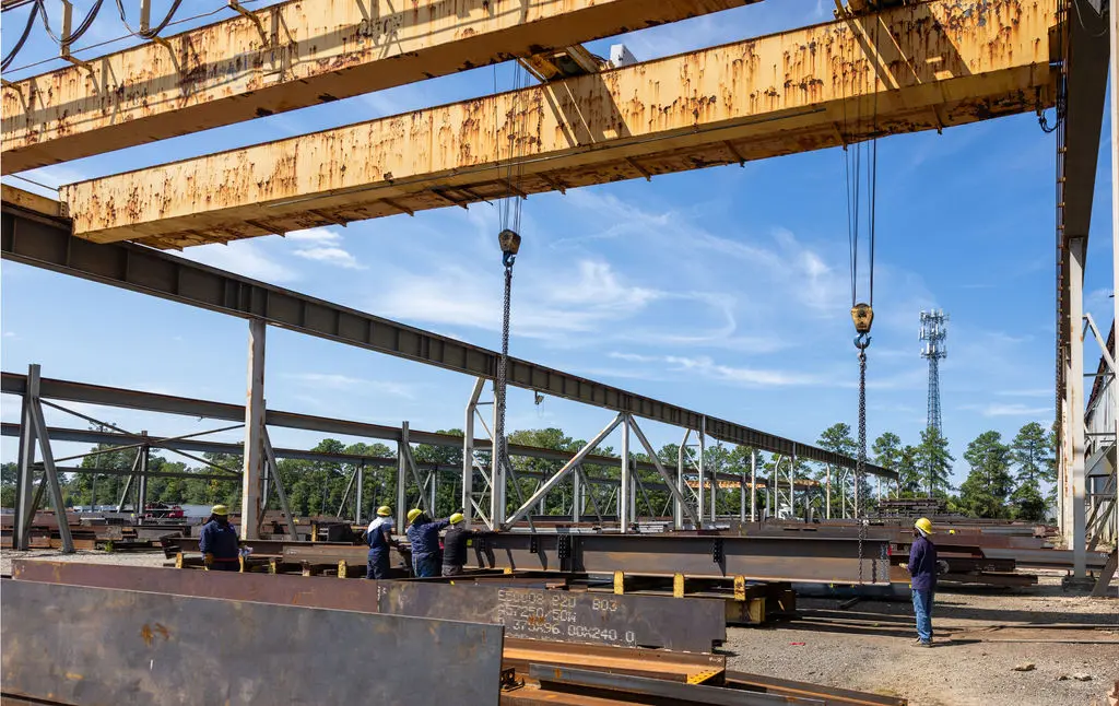 Construction workers guide structural steel beams with overhead crane at outdoor fabrication yard