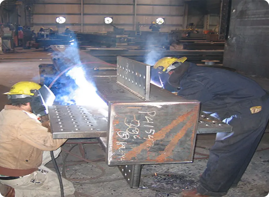 Two welders fabricate a structural steel beam in a metal shop, sparks and smoke during industrial welding.