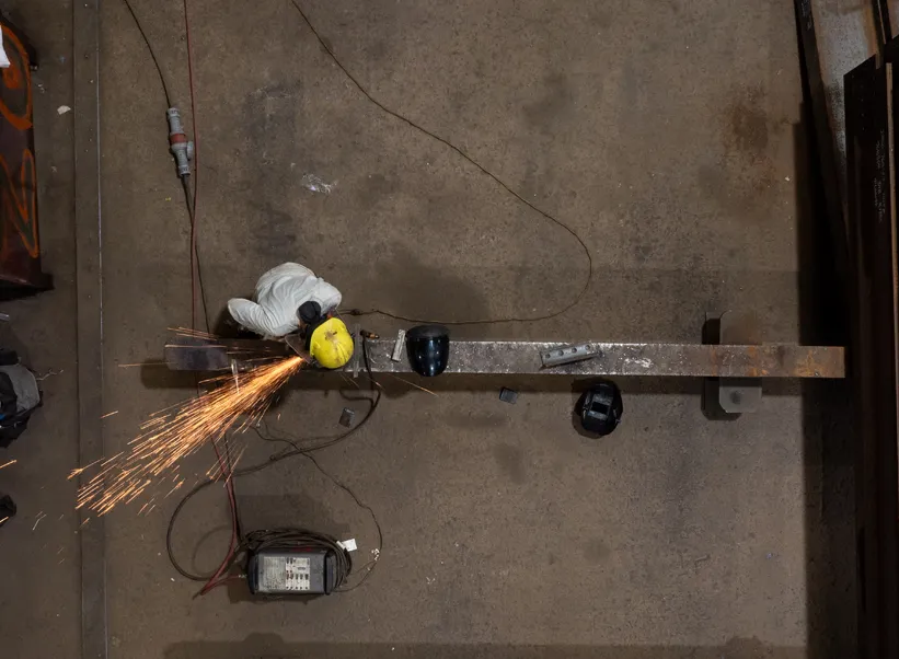 Overhead view of metalworker grinding a steel beam with an angle grinder, sparks flying in an industrial workshop.