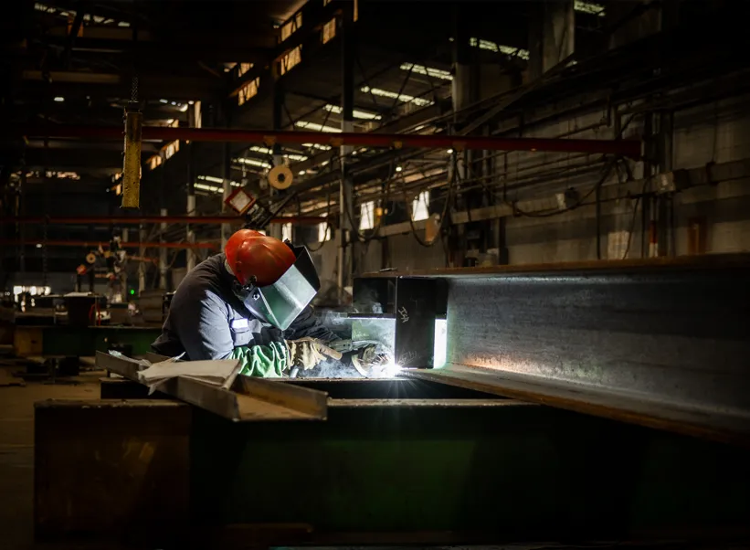 Industrial welder in protective gear welding a steel beam in a manufacturing plant, sparks flying in dark workshop.
