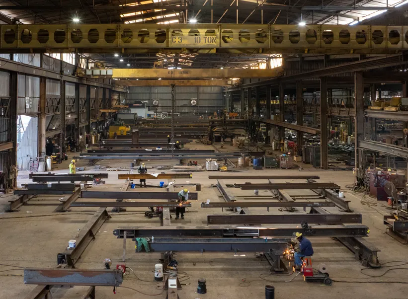 Steel fabrication shop with workers welding beams under 10-ton overhead cranes in a large industrial warehouse.
